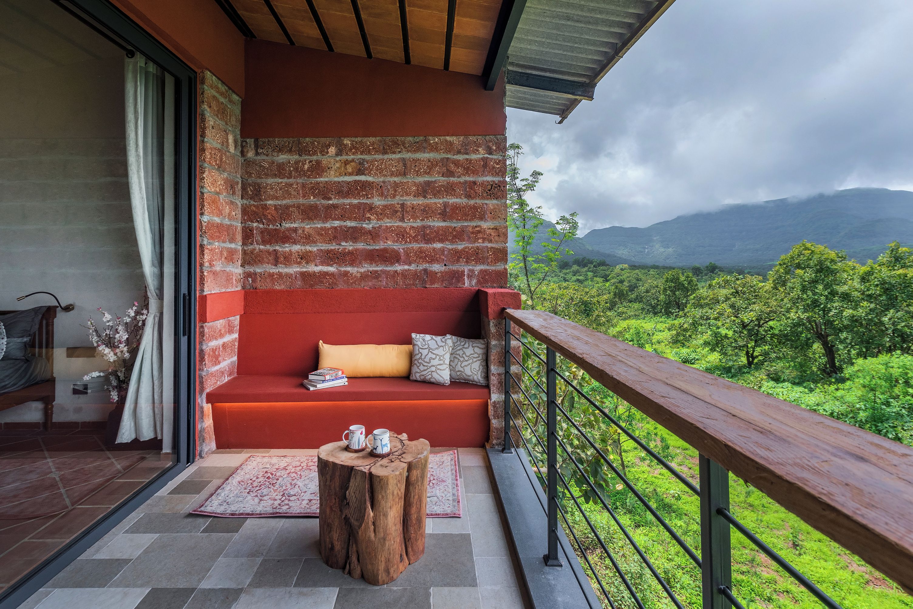 Un balcón con un sofá rojo, un taburete de madera y vistas a exuberante follaje y montañas.