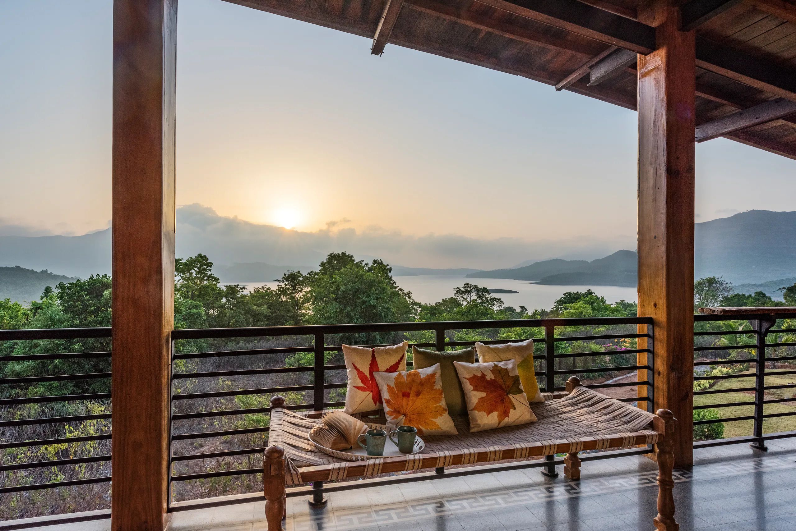 A wooden deck with a bench and cushions, overlooking a lake and mountains.