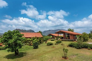 A two-story brick house with a red roof, surrounded by lush greenery and mountains in the background. - Satori Mulshi (Paud)