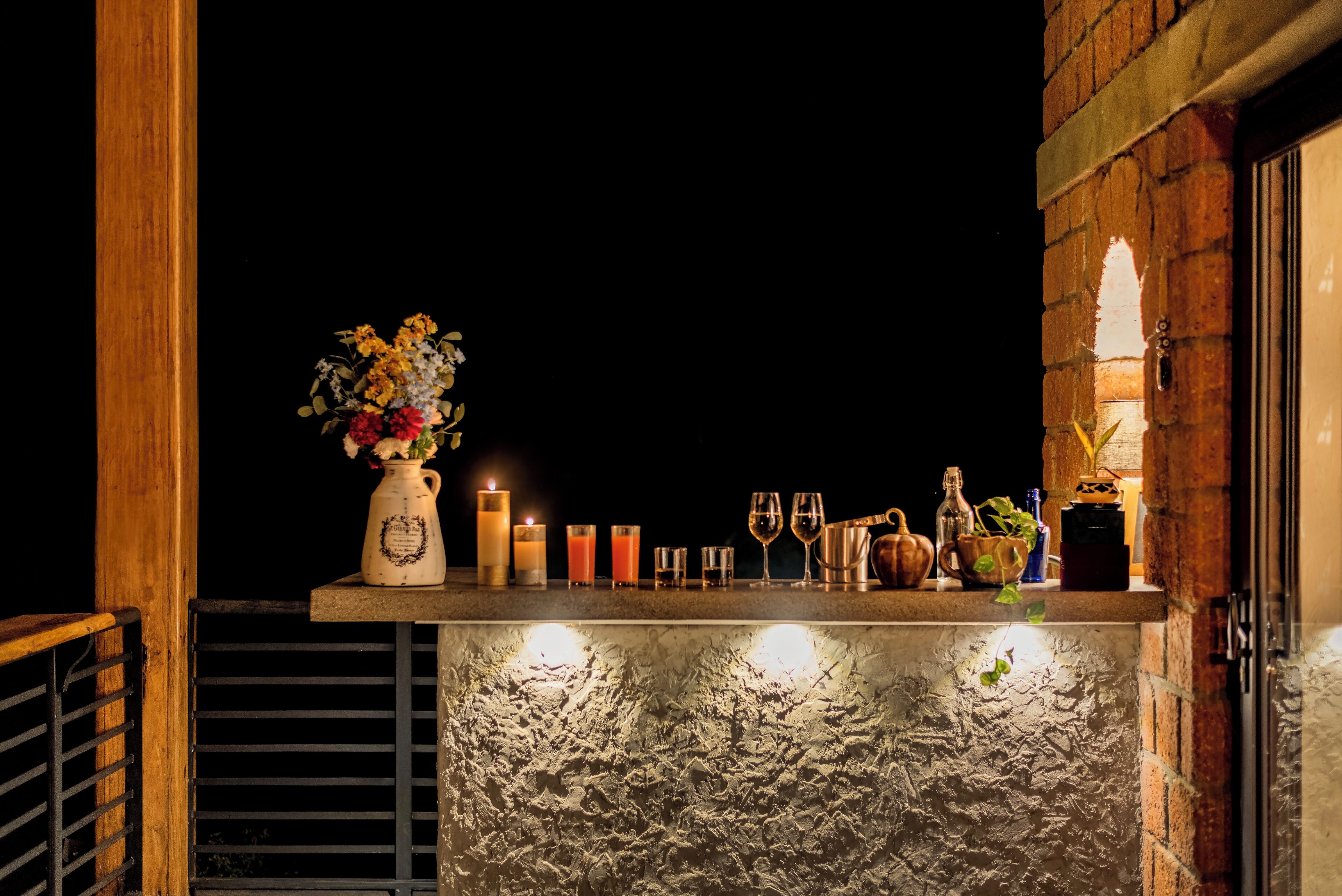 A bar area with a rustic brick wall, a wooden railing, and a shelf with candles, glasses, and a vase of flowers.