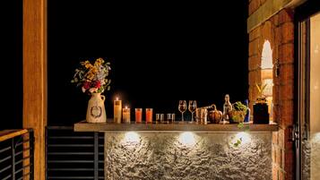 A bar area with a rustic brick wall, a wooden railing, and a shelf with candles, glasses, and a vase of flowers.