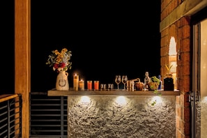 A bar area with a rustic brick wall, a wooden railing, and a shelf with candles, glasses, and a vase of flowers.