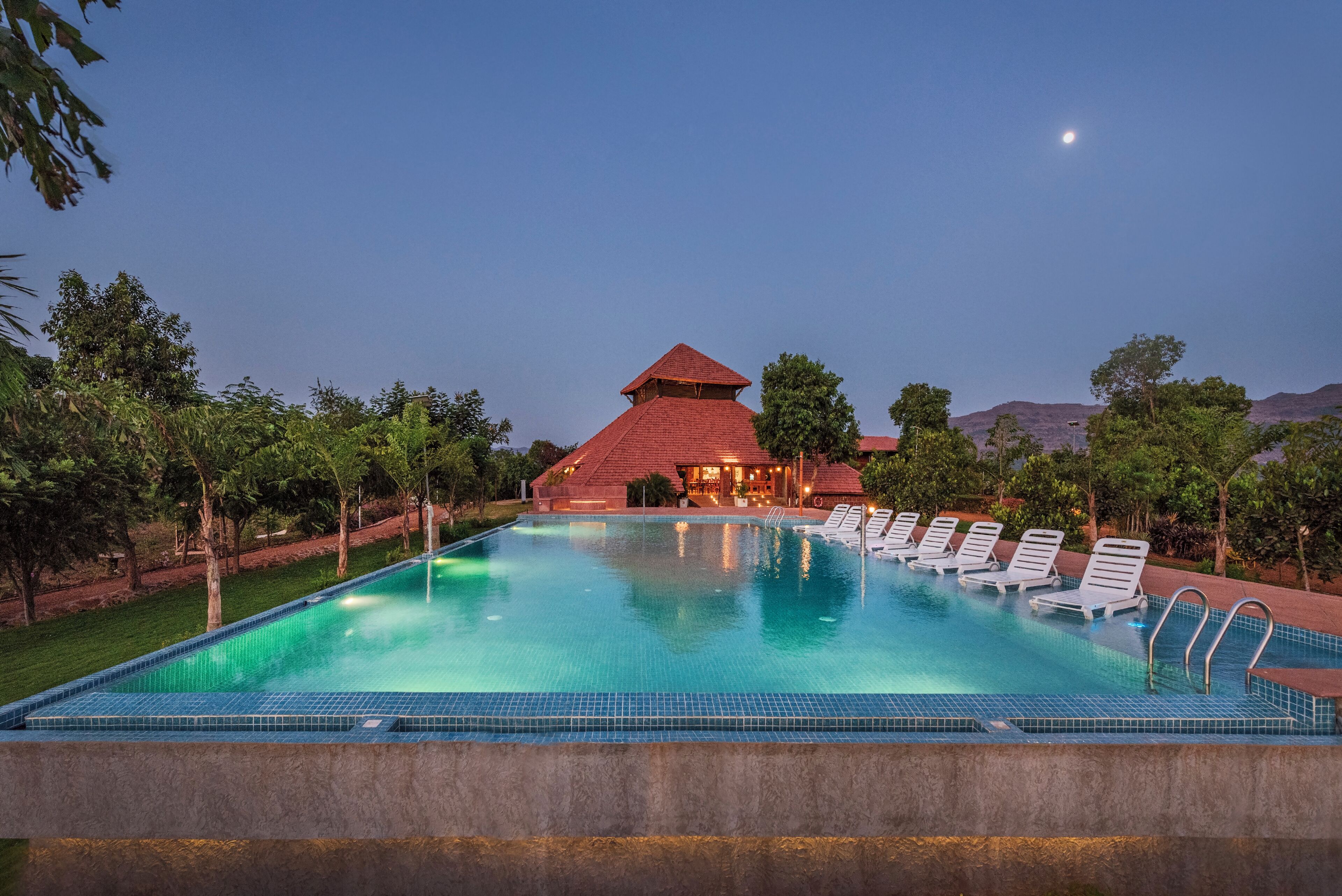A swimming pool with lounge chairs, a building with a red roof, and trees in the background.