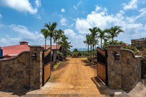 A dirt path leading to a gated entrance with stone walls and a red-roofed building. - Satori Mulshi (Paud)