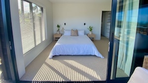 Een slaapkamer met een groot bed, twee nachtkastjes, een raam met jaloezieën en een deur die naar een andere kamer leidt.