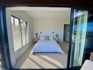 Ein Schlafzimmer mit einem großen Bett, zwei Nachttischen, einem Fenster mit Jalousien und einer Tür zu einem anderen Raum.