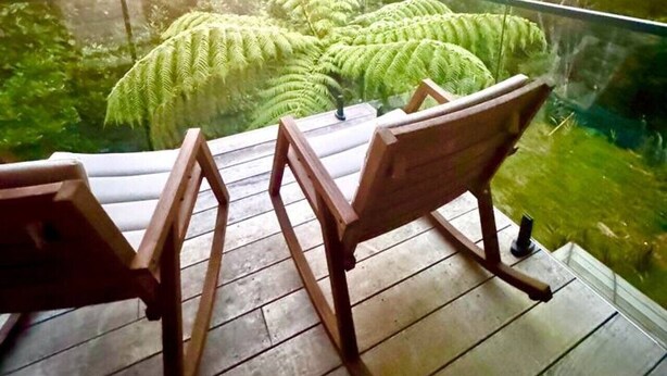 Een balkon met twee houten stoelen, gericht op een weelderig groen landschap met varens en een berg op de achtergrond.