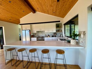 A modern kitchen with white cabinets, a wooden ceiling, and a bar area with stools. - Nestled in Nature•3 Bedroom•Spa Pool•Sauna (Raglan)