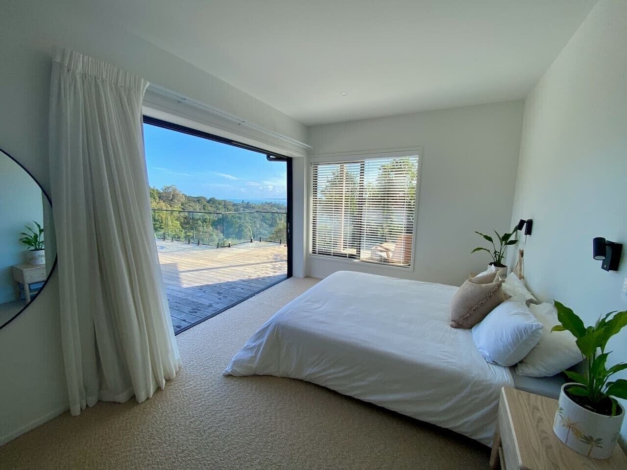 A bedroom with a large bed, a window with blinds, a potted plant, and a view of a balcony.