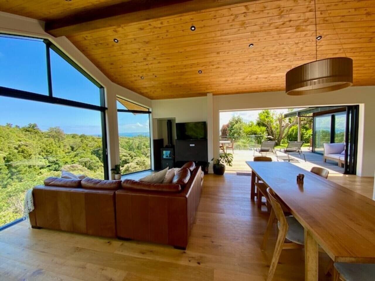 A modern living room with a wooden ceiling, a large window, a sofa, a TV, and a dining area with a table and chairs.
