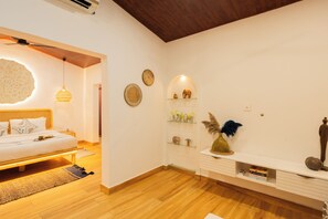 A modern bedroom with a wooden floor, a bed with white bedding, a bedside table, a wall-mounted shelf with decorative items, and a ceiling fan.