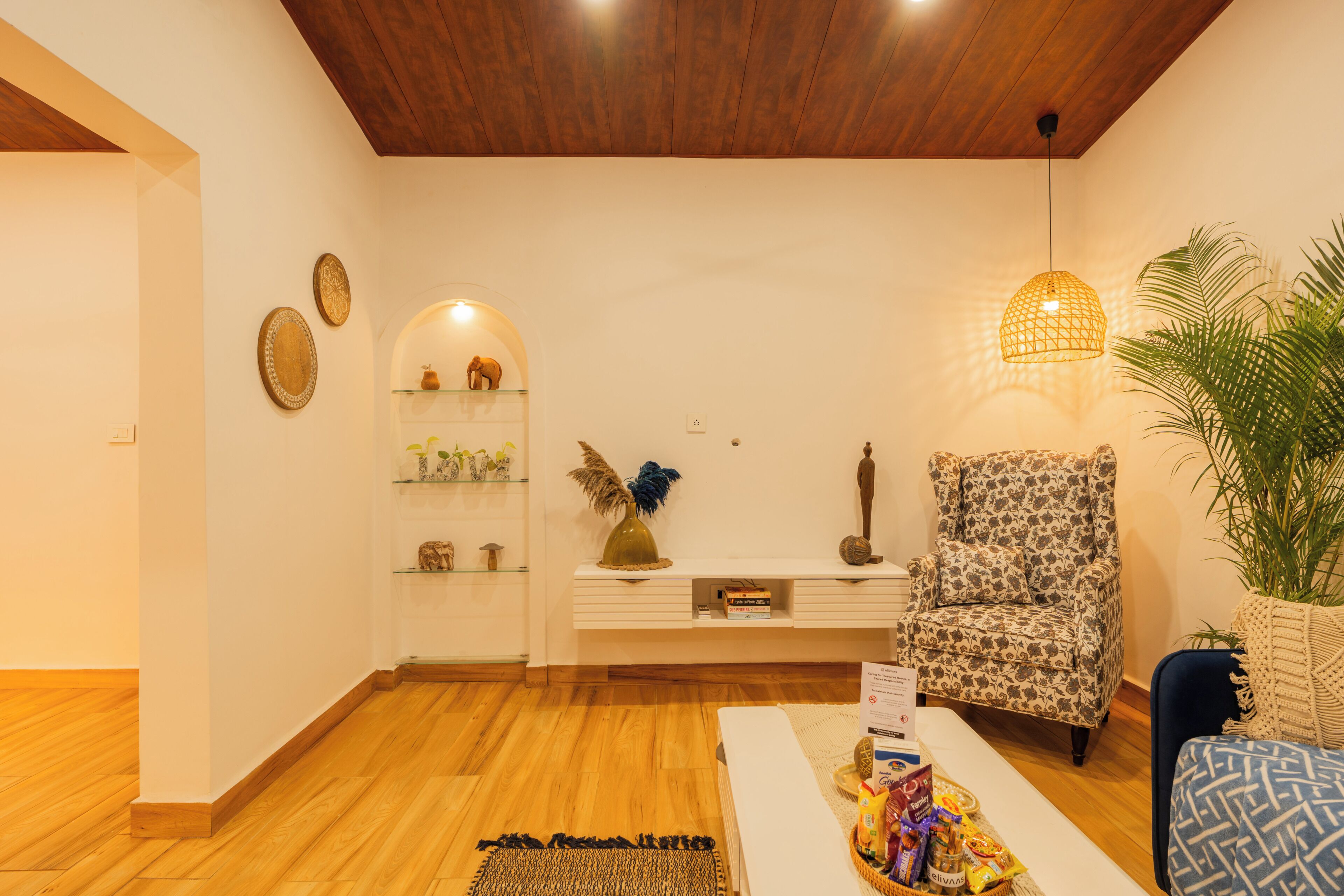 A living room with a wooden ceiling, a patterned armchair, a white sofa, a coffee table with snacks, and a shelf with decorative items.