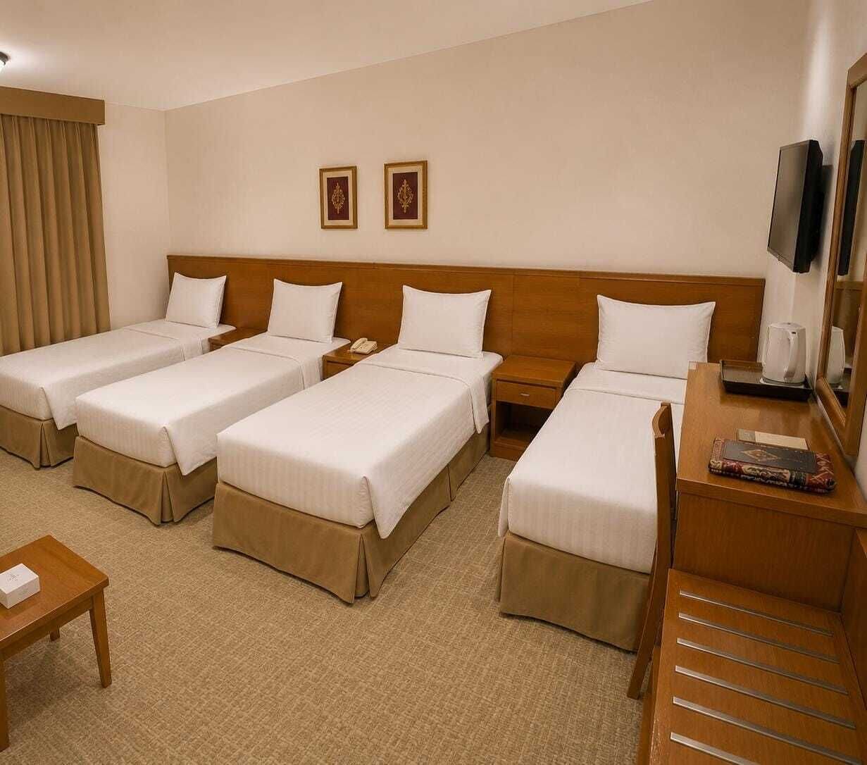 Een hotelkamer met vier eenpersoonsbedden, een houten hoofdeinde, een bureau met een stoel, een televisie en een klein tafeltje met een boek.