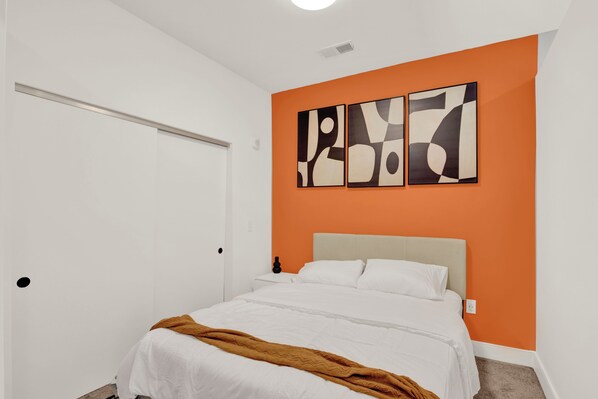 A bedroom with a bed, a white door, and an orange accent wall with abstract art. - Cozy 1BR OTR Suite | Pool, Gym & City Views (Cincinnati)