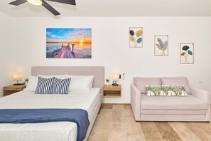 Ein Schlafzimmer mit einem Bett, einem Sofa, einem Nachttisch mit Lampe und einem Wandbild, das einen Sonnenuntergang über einem Steg zeigt.