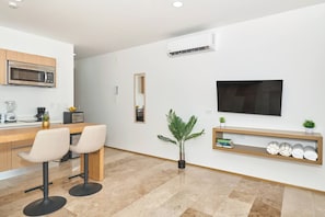 Eine moderne Küche mit Mikrowelle, Mixer und Kaffeemaschine. Ein Essbereich mit zwei Stühlen und einem kleinen Tisch ist vorhanden. Ein Fernseher ist an der Wand befestigt, und eine Pflanze steht auf dem Boden.