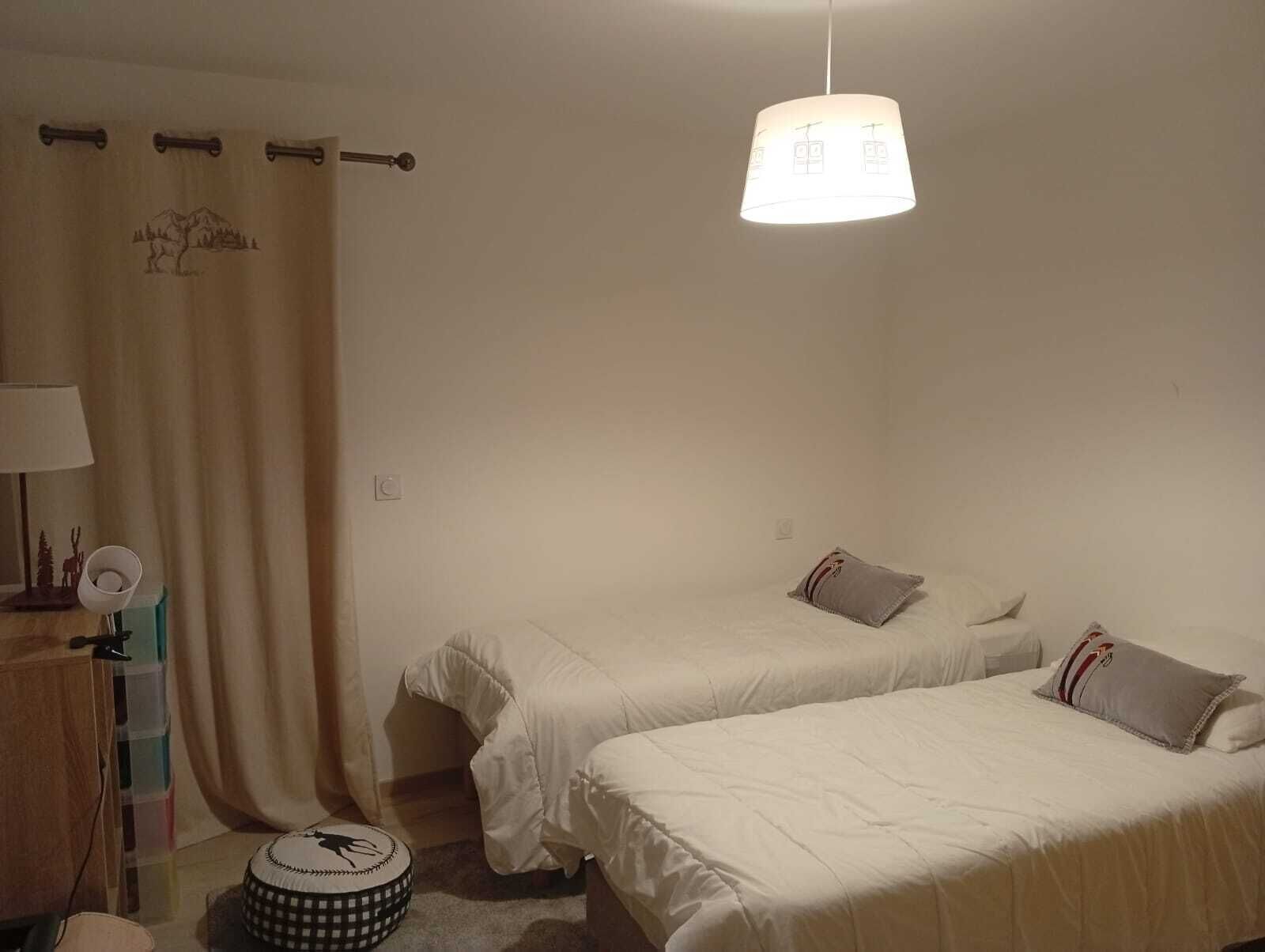 A bedroom with two beds, a hanging light fixture, a curtain with a mountain design, and a small round table with a lamp.