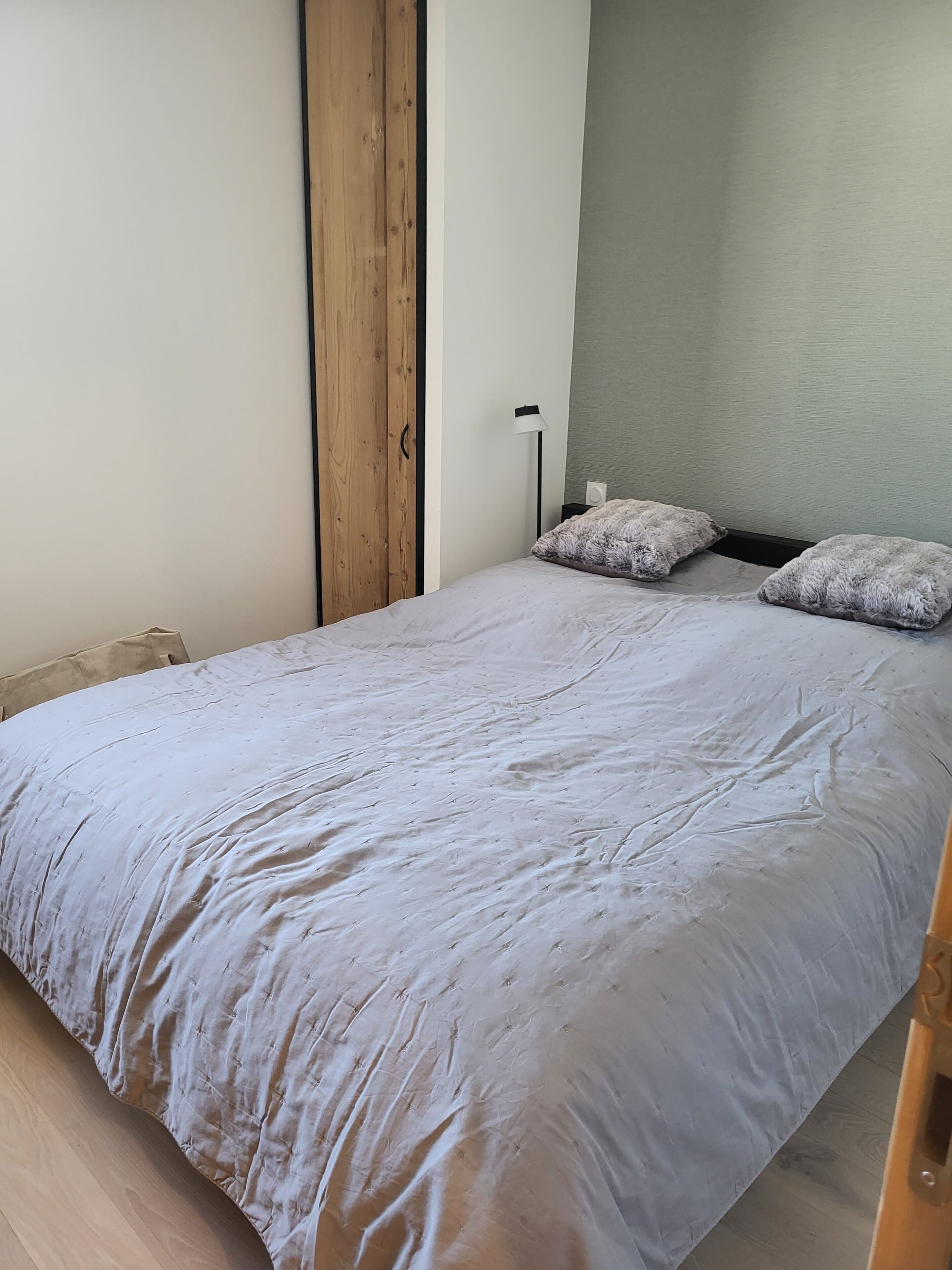 Un lit simple avec une couette grise, une armoire en bois et une lampe de chevet.