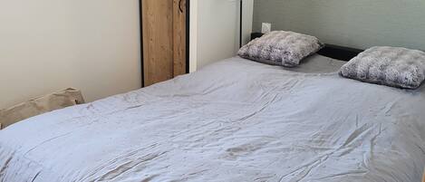 Un lit simple avec une couette grise, une armoire en bois et une lampe de chevet.