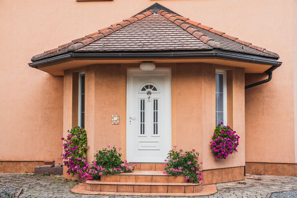 Een huis met een witte deur, een pannendak en roze bloemen rondom de ingang.