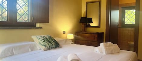 A hotel room with a bed, a bedside lamp, a mirror, and a window with a view of greenery.
