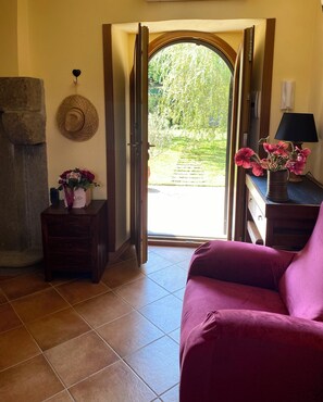 A room with a wooden door leading to a garden, a stone wall, a small table with a vase of flowers, and a purple sofa.