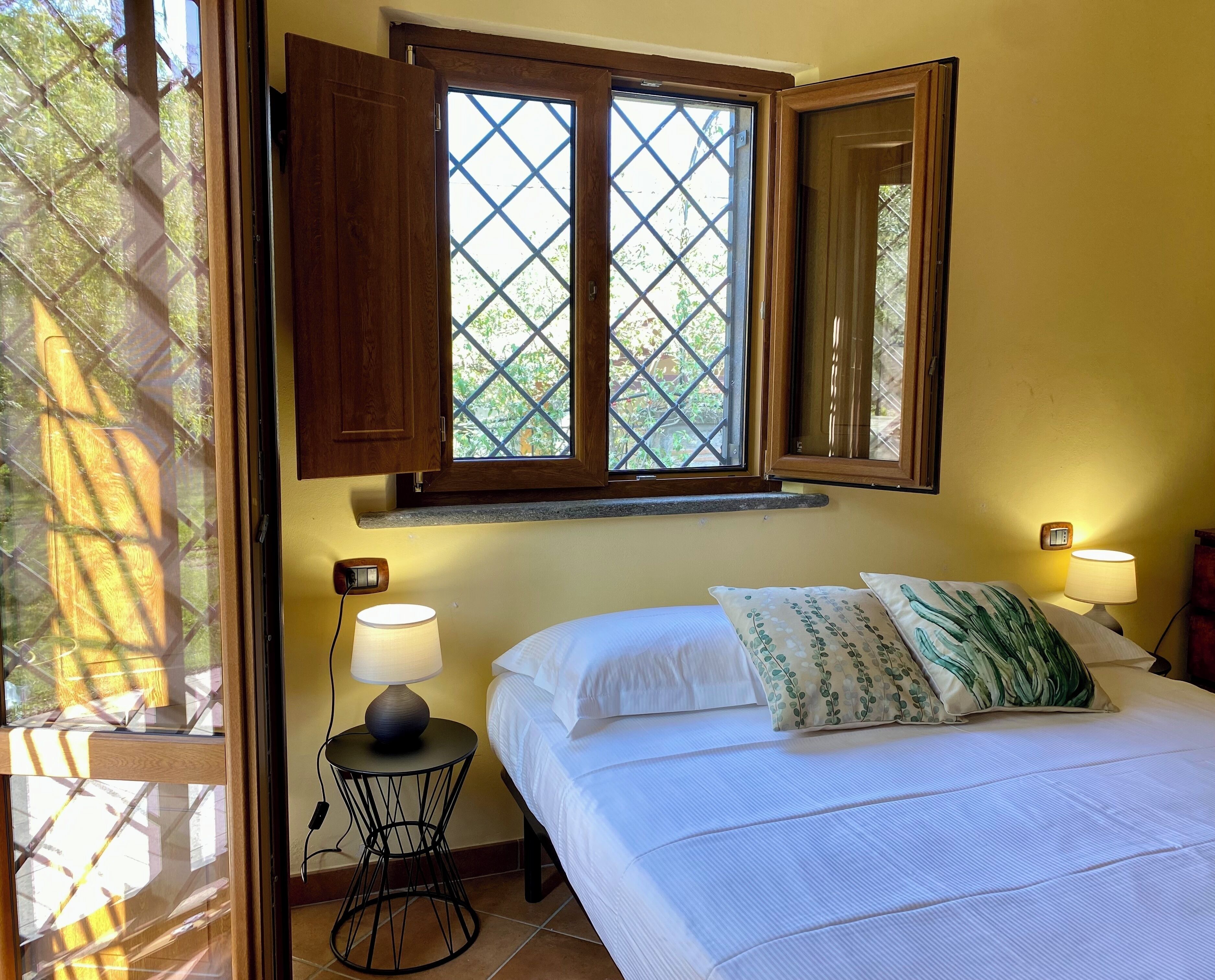 Un dormitorio con una cama, dos lámparas de noche, una mesita de noche, una ventana con contraventanas de madera y un termostato fijado a la pared.