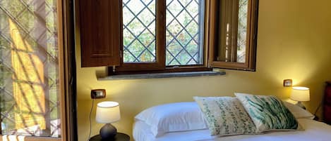 Un dormitorio con una cama, dos lámparas de noche, una mesita de noche, una ventana con contraventanas de madera y un termostato fijado a la pared.