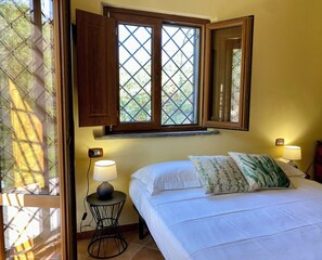Ein Schlafzimmer mit einem Bett, zwei Nachttischlampen, einem Nachttisch, einem Fenster mit Holzjalousien und einem an der Wand montierten Thermostat.