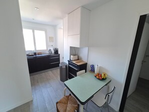 A compact kitchen with a dining table, a trash bin, and a window providing natural light. - Comfortable apartment near the beach - 5 guests (Les Sables-d'Olonne)