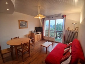 Une pièce dotée d’un plancher en bois, d’une table à manger avec des chaises, d’un canapé, d’un téléviseur posé sur un meuble et d’un balcon offrant une vue sur la verdure et les montagnes.