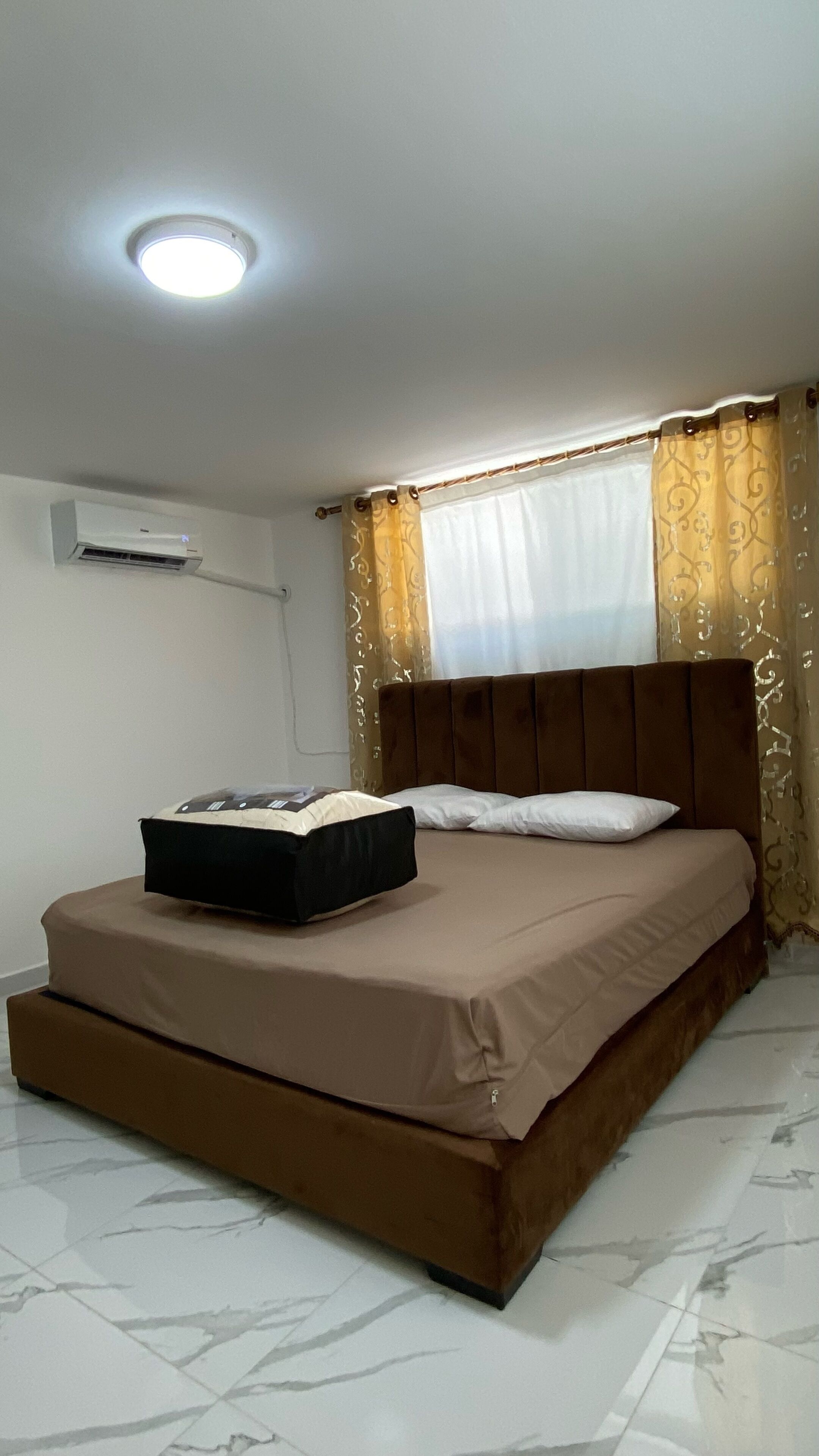 Ein Schlafzimmer mit Bett, Nachttisch, Wand-Klimaanlage und einem Fenster mit Vorhängen.