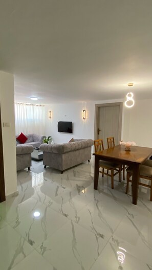 A living room with a sofa, a dining table with chairs, a flat-screen TV mounted on the wall, and a decorative light fixture.