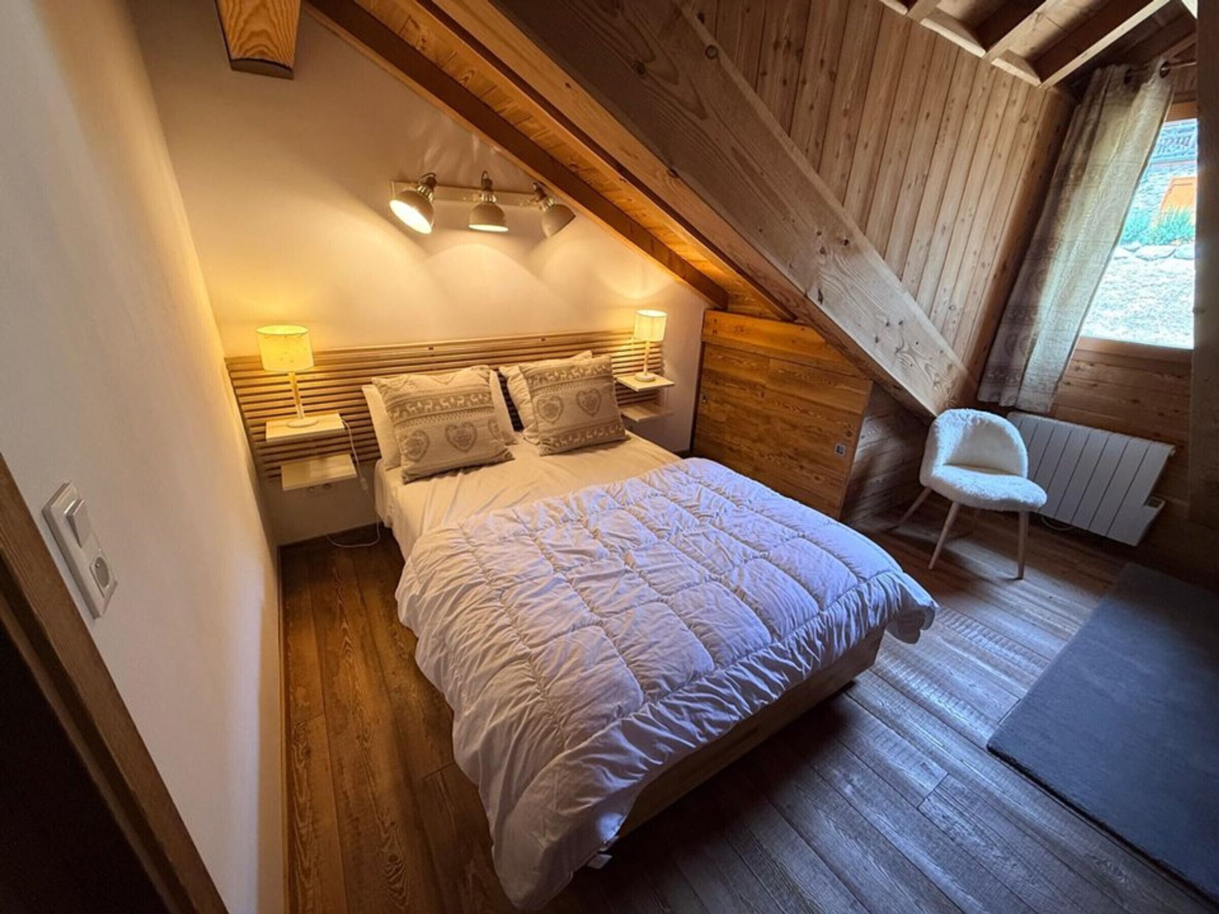 A bedroom with a sloped ceiling, a bed with a quilt, bedside lamps, and a chair.