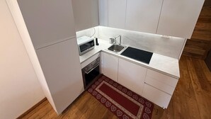 Ett kompakt kök med vita skåp, en inbyggd mikrovågsugn, en diskho och en röd, mönstrad matta.