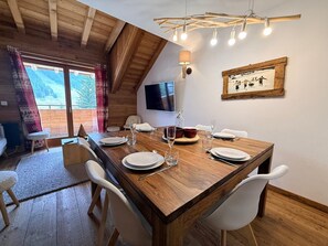 Ein Essbereich mit einem Holztisch, gedeckt für vier Personen, weißen Stühlen, einem Fernseher an der Wand und einem gerahmten Bild.
