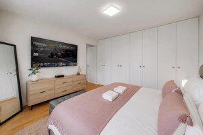 Un dormitori modern amb un llit gran, una televisió penjada a la paret, un taulell de nit de fusta i un armari.