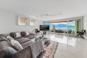 Uma sala de estar moderna com um conjunto de sofás, uma mesa de centro e uma televisão. Há uma varanda com vista para as montanhas.