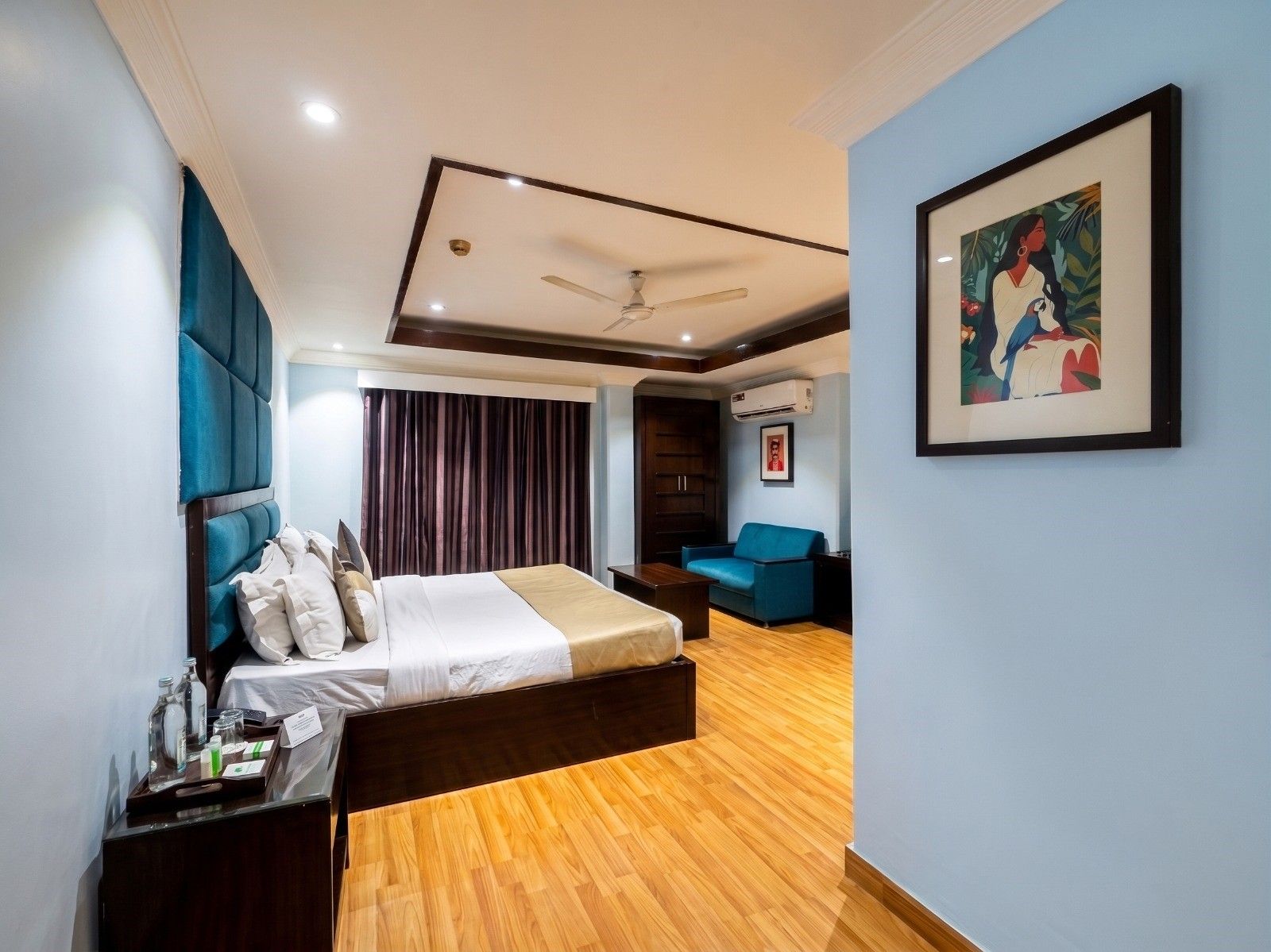 A modern hotel room with a bed, a desk with a tray of toiletries, a blue sofa, and a ceiling fan.