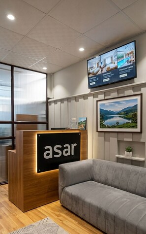 Un área de recepción con un escritorio de madera identificado con 'asar', un sofá gris, un televisor mostrando opciones de habitaciones y un cuadro enmarcado con un paisaje.