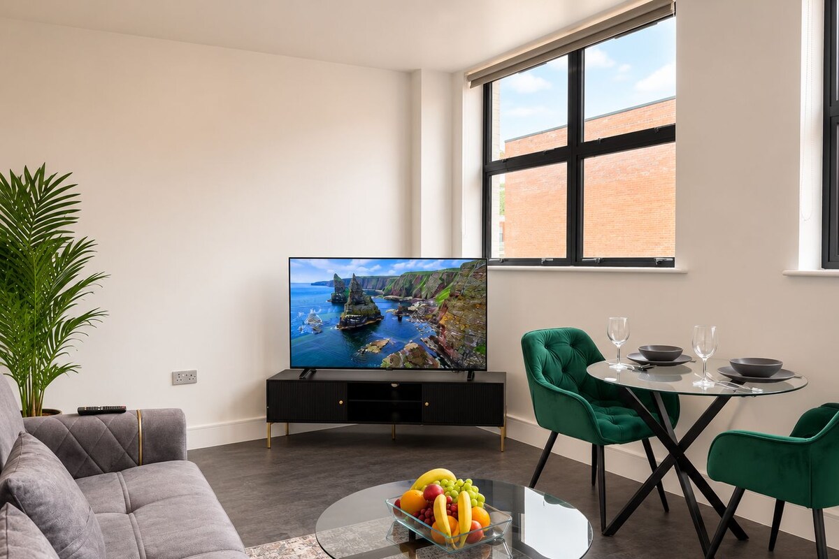 A modern living room with a flat-screen TV, a sofa, a glass coffee table with fruit, and a dining area with chairs and a table.