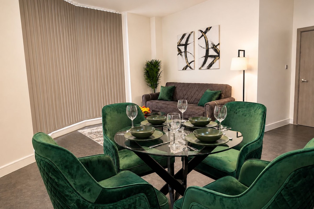 A dining area with a round glass table, green upholstered chairs, and a brown sofa in the background.