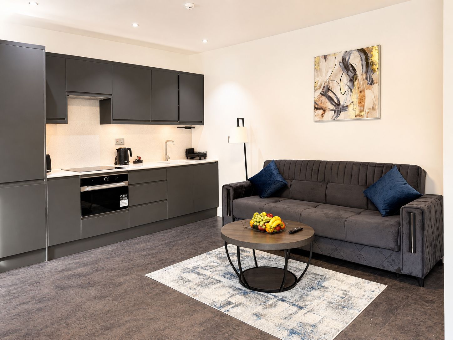 Eine moderne Küche mit dunklen Schränken, ein Essbereich mit grauem Sofa und blauen Kissen sowie eine Obstschale auf einem runden Tisch.