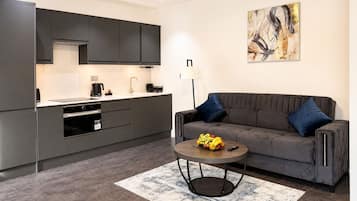 Eine moderne Küche mit dunklen Schränken, ein Essbereich mit grauem Sofa und blauen Kissen sowie eine Obstschale auf einem runden Tisch.