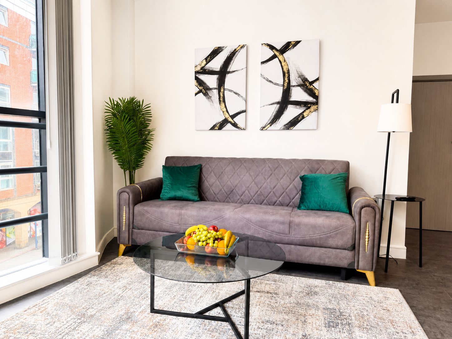 A modern living room with a grey sofa, a glass coffee table with fruit, abstract wall art, a potted plant, and a floor lamp.