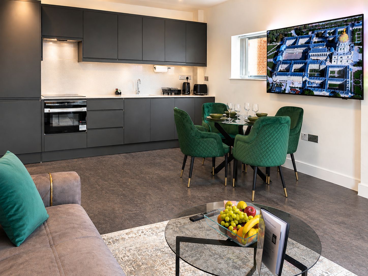 A modern kitchen with dark cabinets, a dining area with green chairs, and a large flat-screen TV on the wall.
