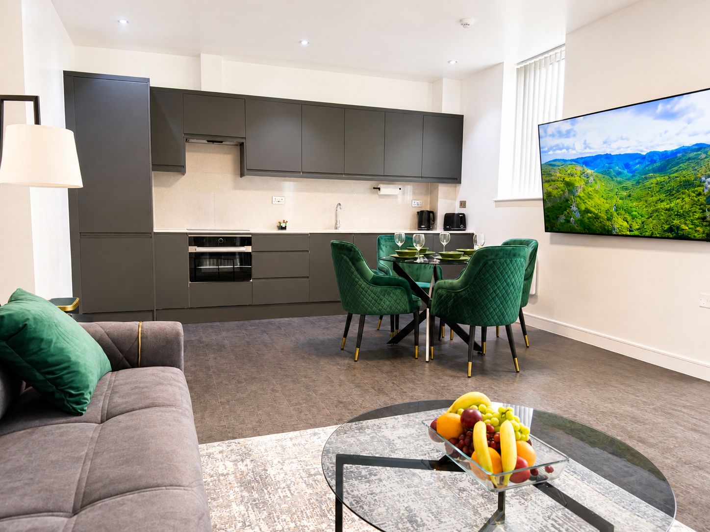 A modern living room with a grey sofa, a glass coffee table with fruit, and a dining area with green chairs.