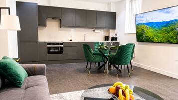 A modern living room with a grey sofa, a glass coffee table with fruit, and a dining area with green chairs.