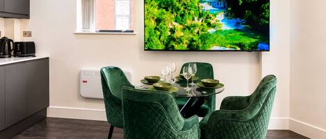 A dining area with a round table, four green chairs, and a flat-screen TV mounted on the wall displaying a nature scene.