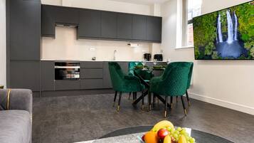 Una cocina moderna con armarios oscuros, un comedor con sillas verdes y un frutero sobre una mesa.